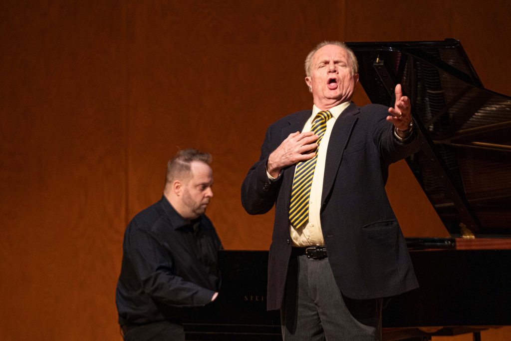 Stephen West sings standing on stage with piano accompanist; with one hand at his chest and the other held out; attired in a dark suit coat and yellow patterned tie.