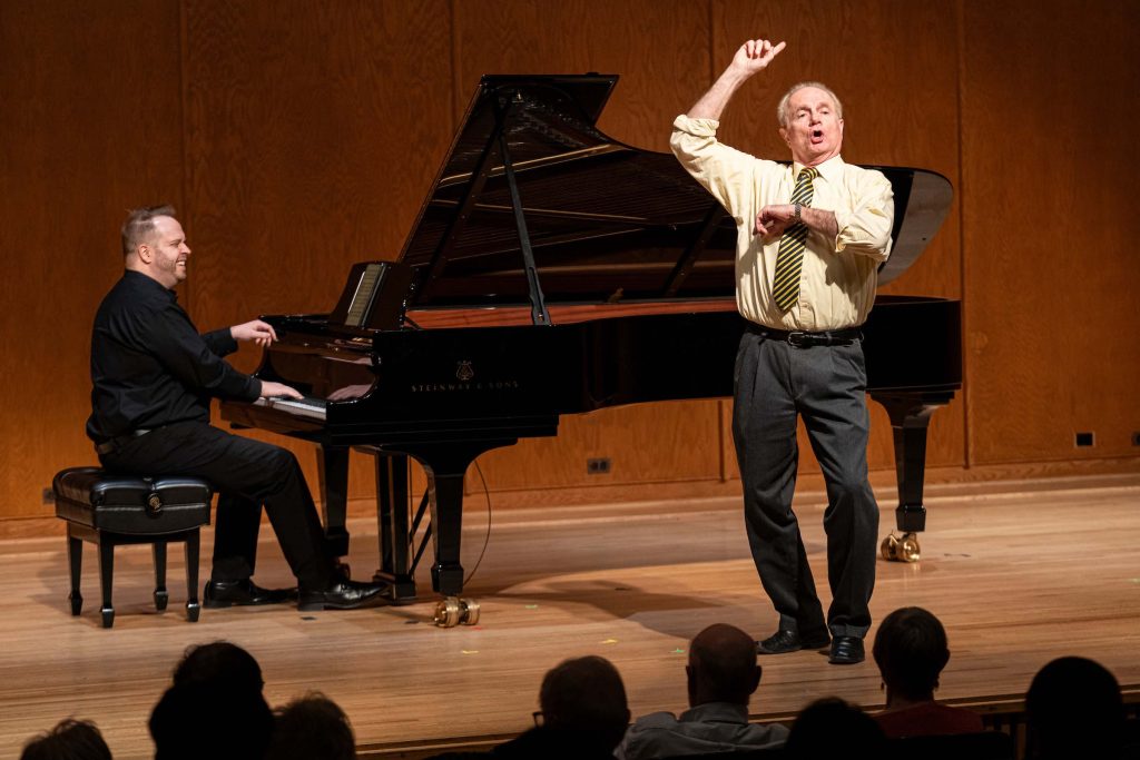 Stephen West sings standing on stage with elbows raised, with a pianist and grand piano in the background and audience heads in the foreground.