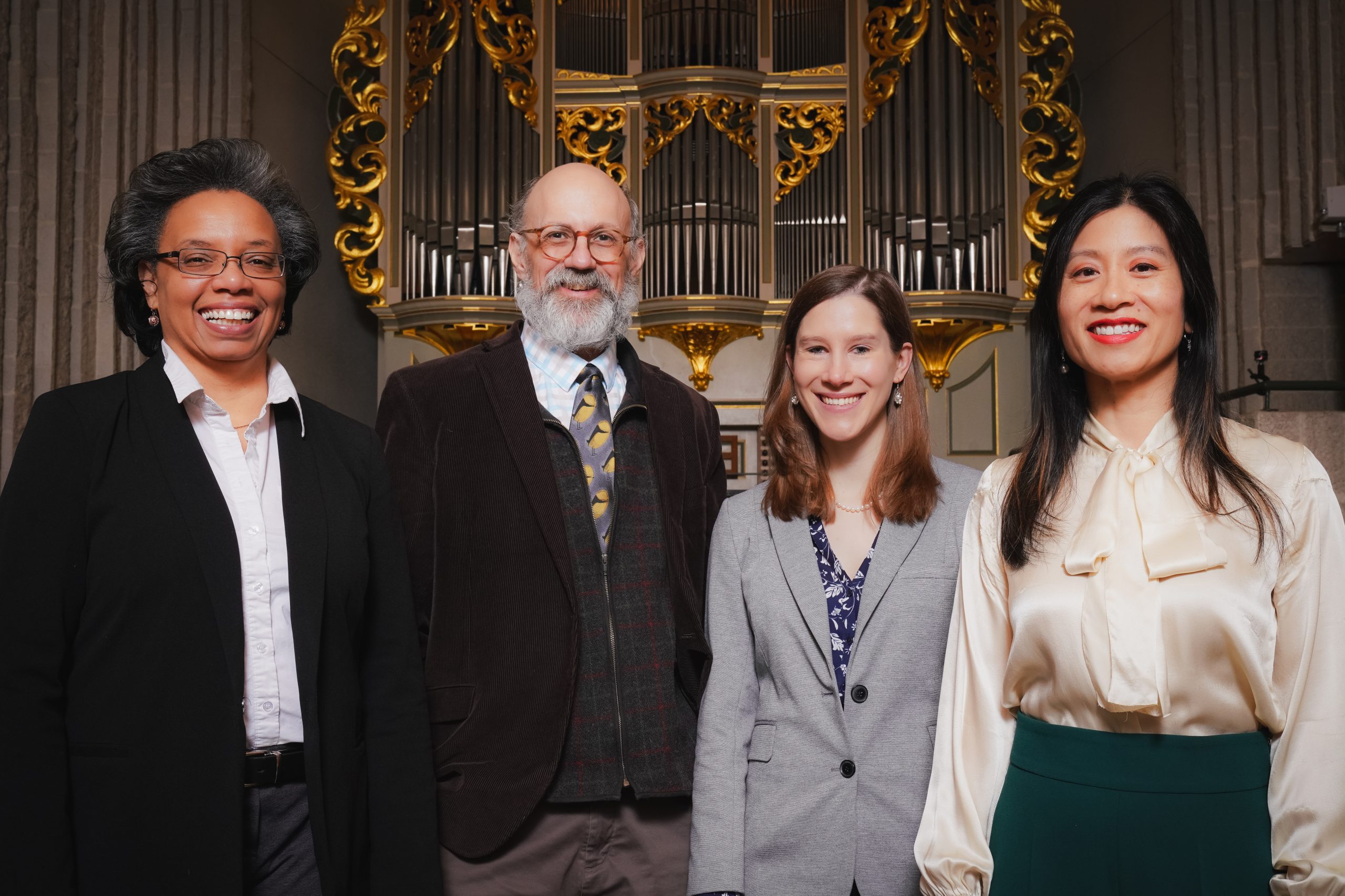 Four people pose standing in business attire, in front of a pipe organ with elaborate gold decoration