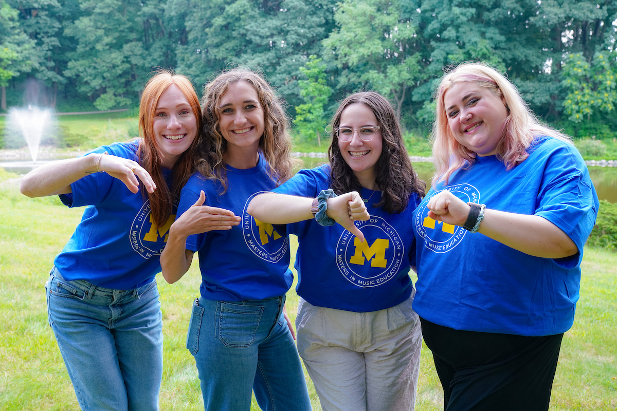 A group of 15 summer masters students post outdoors, holding up their palms as a map of Michigan to show where they are from