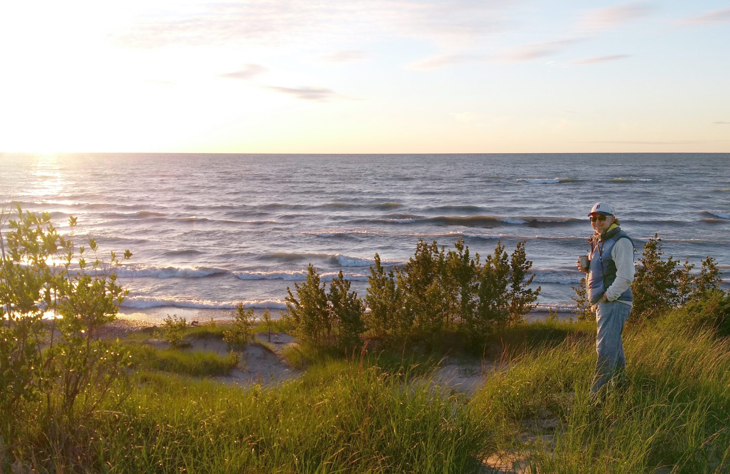 Bill Lucas at sunset on a dune overlooking a major Michigan Lake. He is wearing a Detroit Tigers baseball cap, a puffy vest, sunglasses, and jeans, holding a coffee cup smiling at the camera.