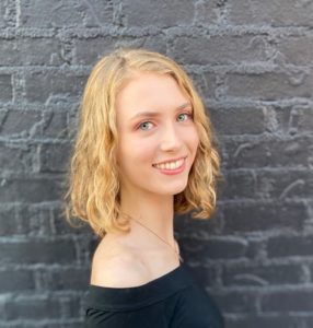 Claire Neidermaier standing in front of a grey brick wall. She has green eyes, shoulder-length wavy blonde hair, is wearing a black off-the-shoulder top, and is smiling towards the camera.
