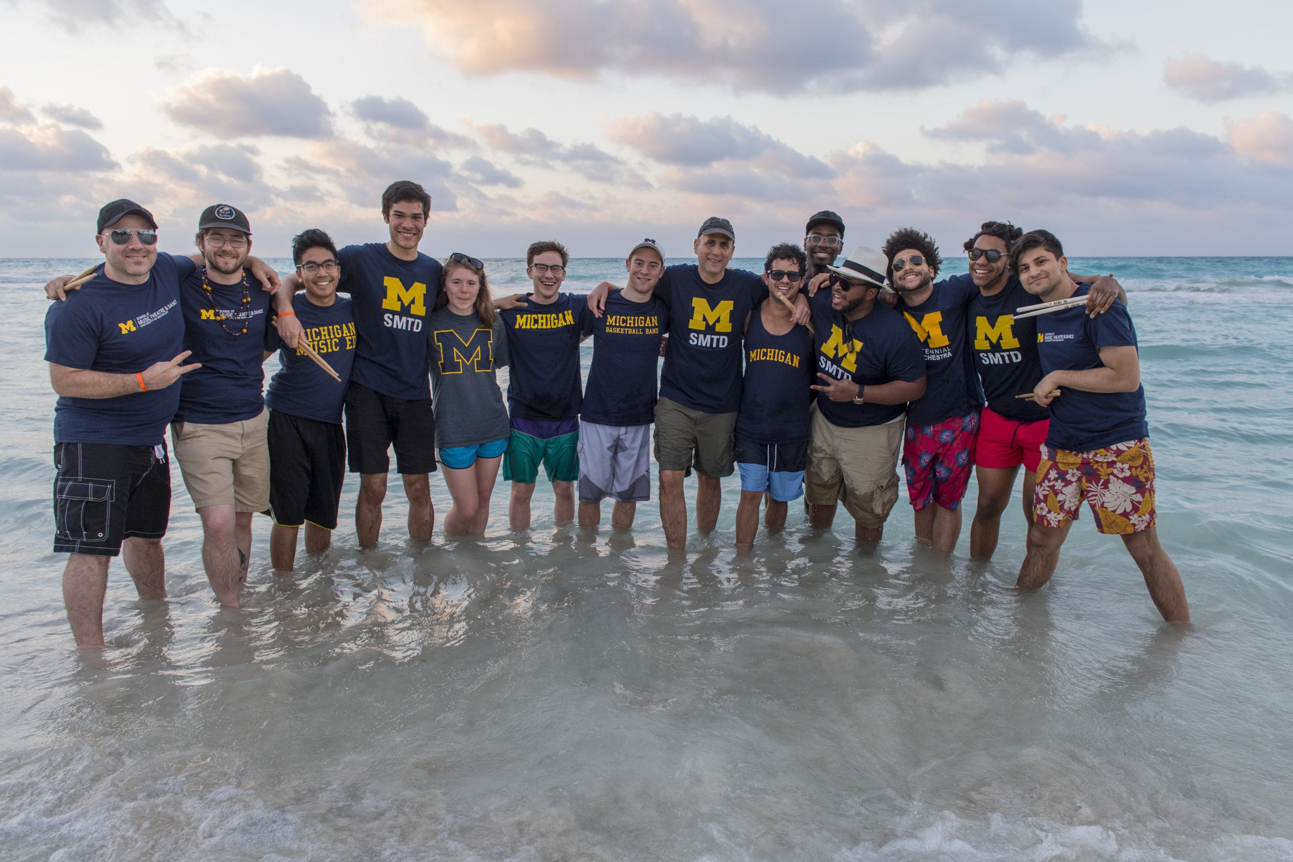 Fourteen SMTD students and faculty wearing various Michigan shirts stand ankle deep in water on a beach in Cuba.