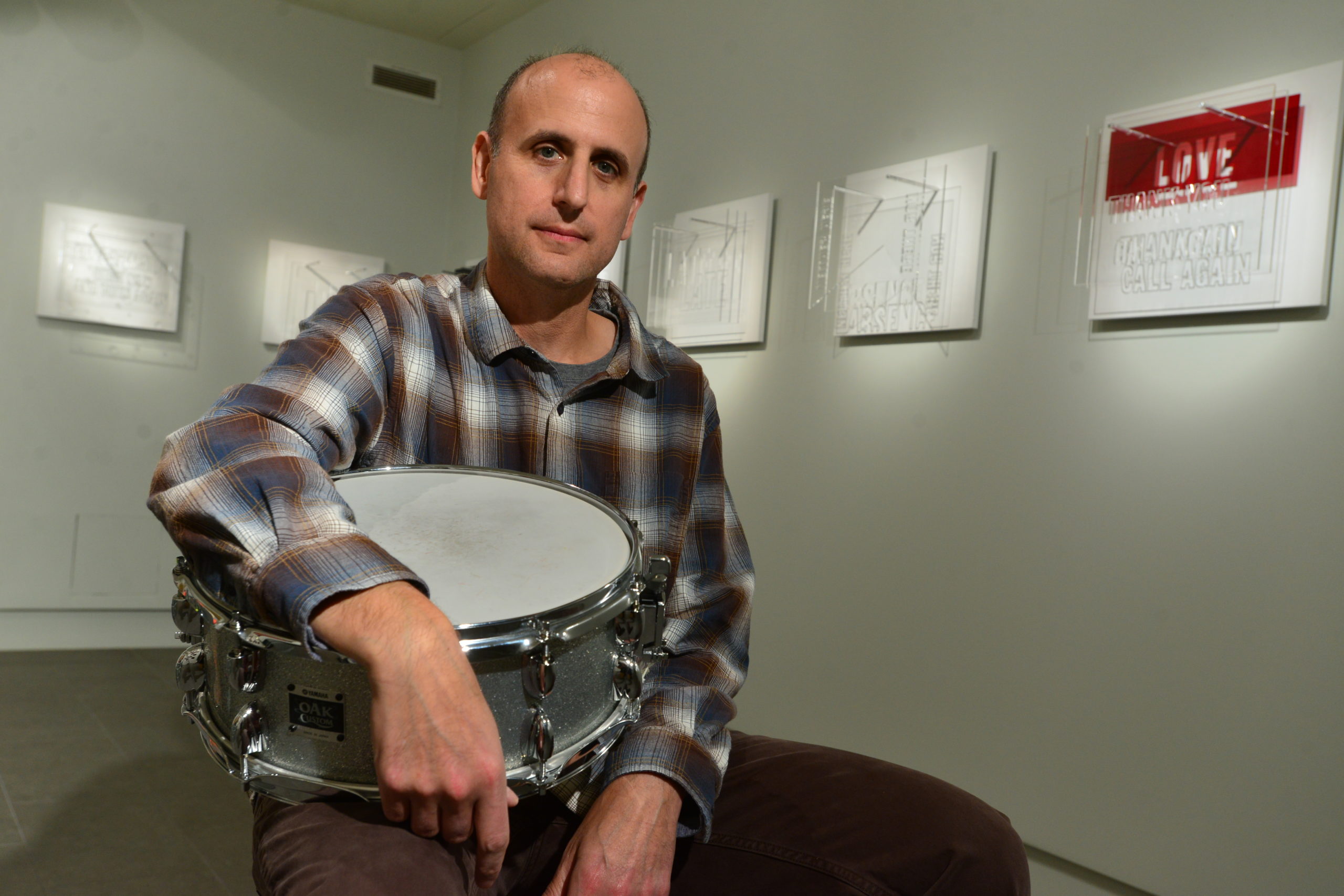 Michael Gould seated in an art gallery with a snare drum in his lap. He is surrounded by an installation of art he has created featuring acrylic cut outs of words on a white background.