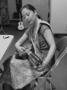 Black and white photo of Christi-Anne Castro in traditional Philippino dress, seated backstage, fixing a string on a banduria, a plucked instrument that is part of the rondalla.