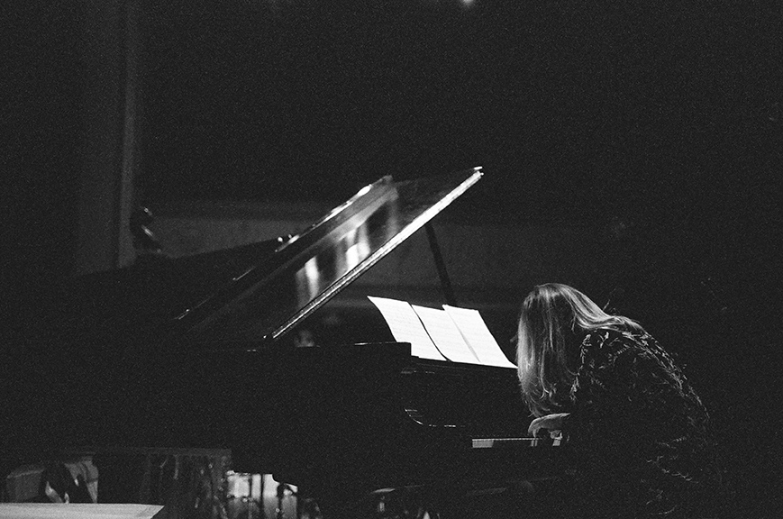 Taken from the back, a black and white photo of Sonya Belaya performing on an open grand piano. Her hair is obscuring her face and she is leaning in towards the keyboard.