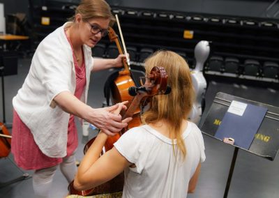 A cello instructor stands working with a seated student on hand positions on the cello.