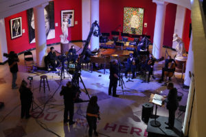The apse of the University of Michigan Museum of Art, showing red walls, large white columns, two marble statues and a modern painting of a person. The floor is covered in cables surrounding the film crew and their equipment as they set up shots of the orchestra for filming.