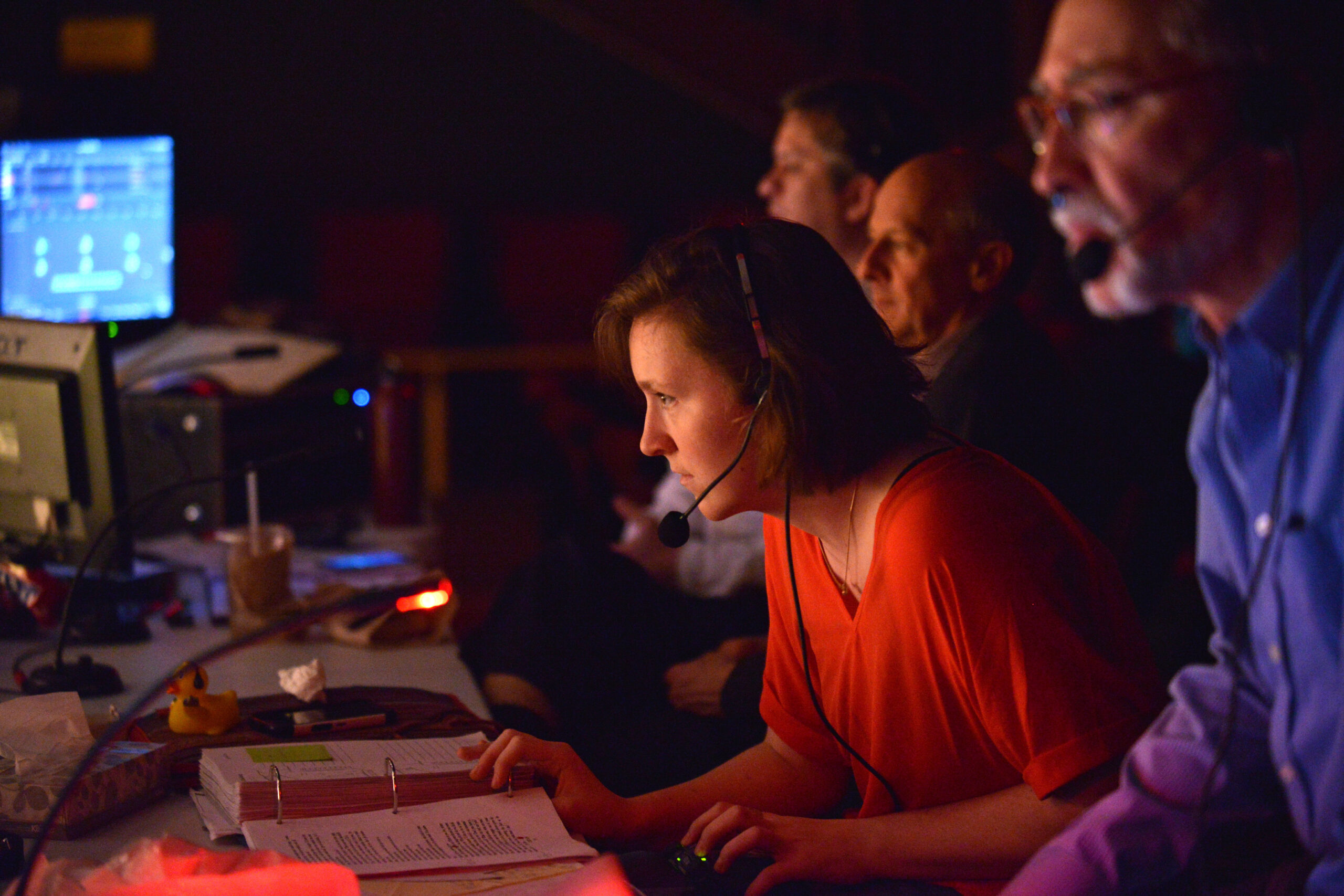 A female student with chin-length hair, wearing a headset, observes a production