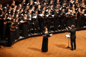 A solo vocalist and conductor stand in front of a large choir performing on stage