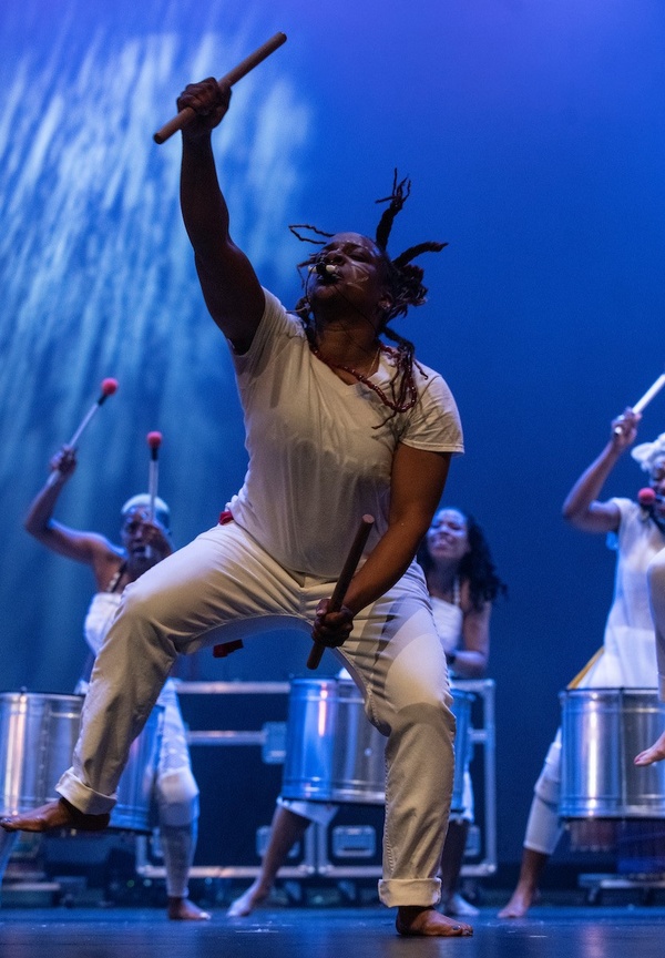 Dancer performs on stage holding percussion mallets, with three drummers in background