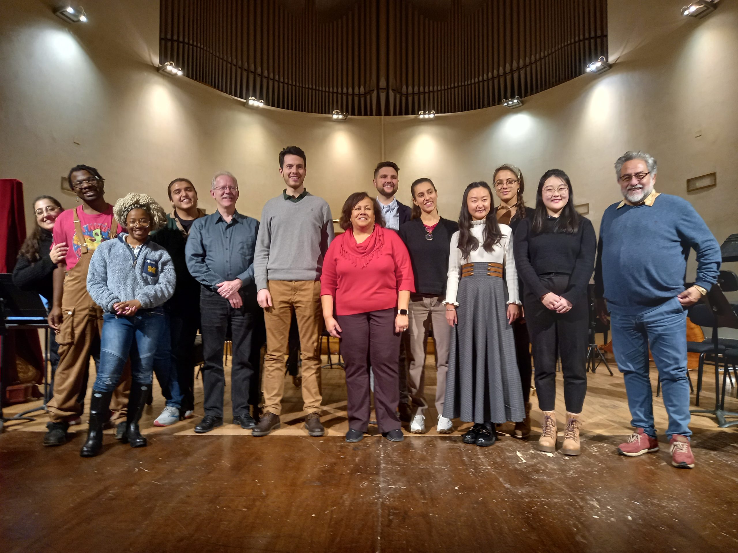 Thirteen individuals pose standing on a stage with backlighting