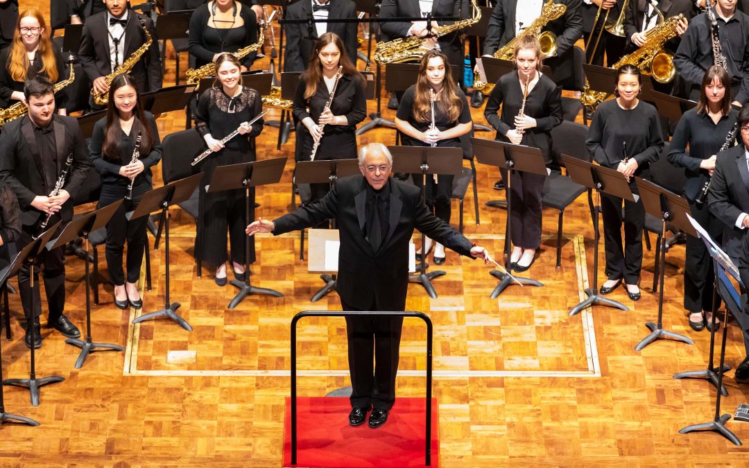 A conductor on stage faces the audience with band members standing behind him.