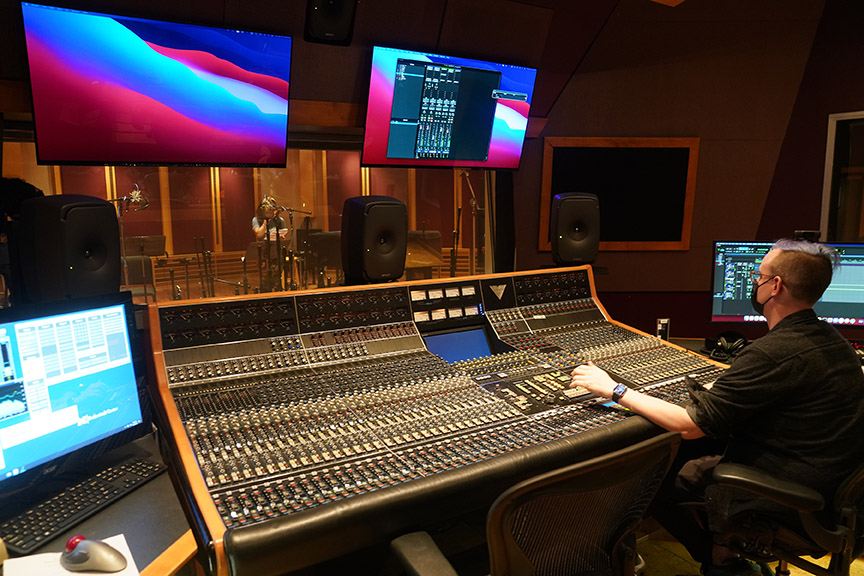 A masked technician works at a large mixing board in a recording studio with four mounted monitor screens; another person is seen inside the recording booth.