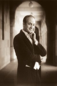 A man poses in formal wear with left hand held to his chin.