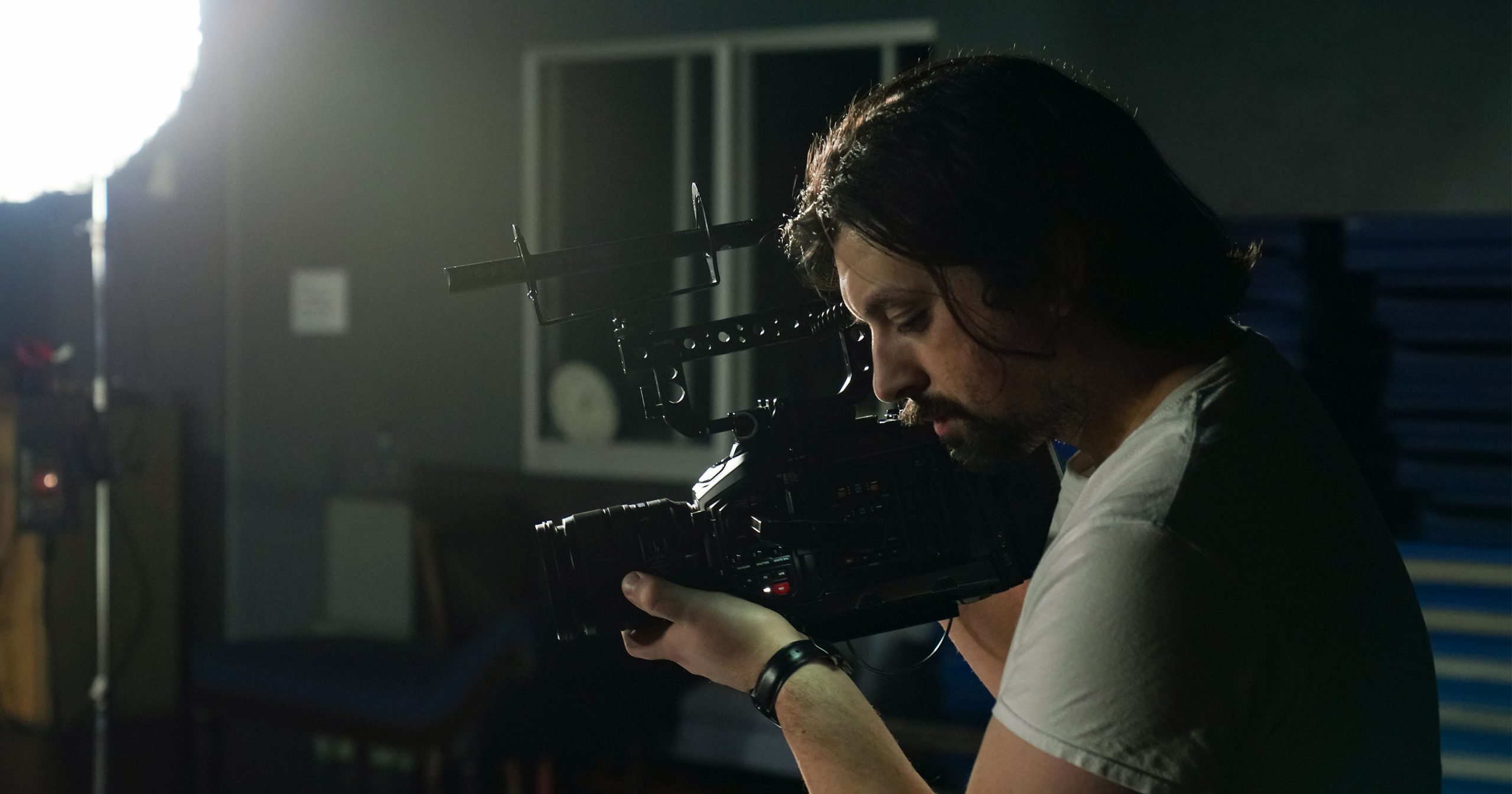 A man films with his eye held to the video camera; a spotlight shines from the upper left corner.