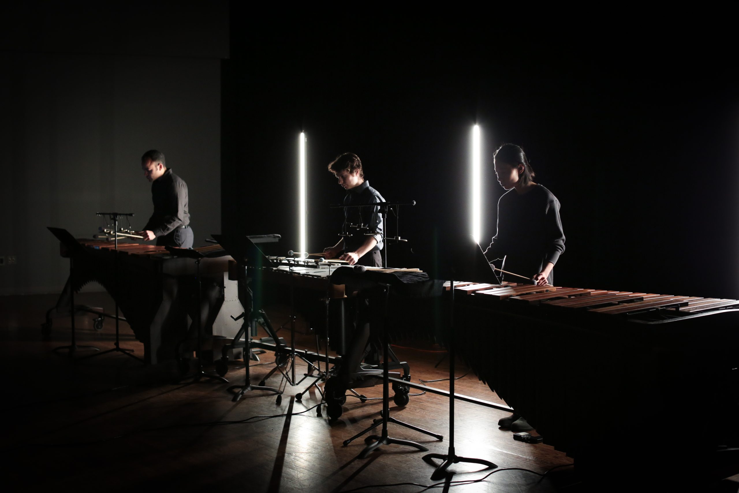 Three individuals perform on xylophones; two column-shaped lights shine from behind them onto a dark and shadowy stage.