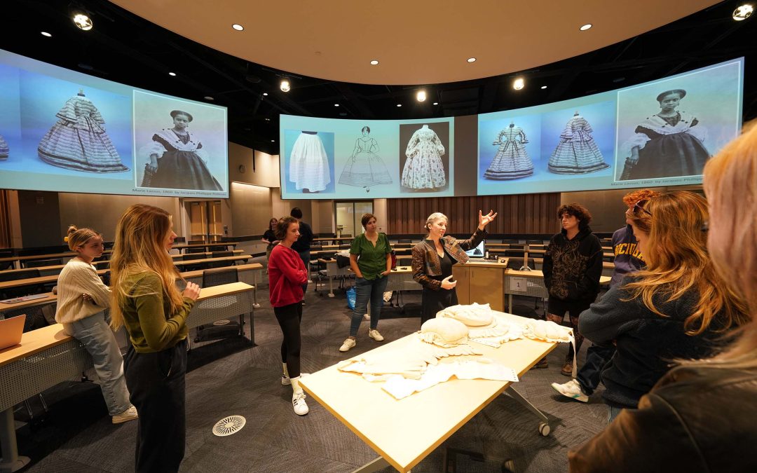 A round classroom with 19th-century garments on screen and on a table; an instructor presents to students while all stand.
