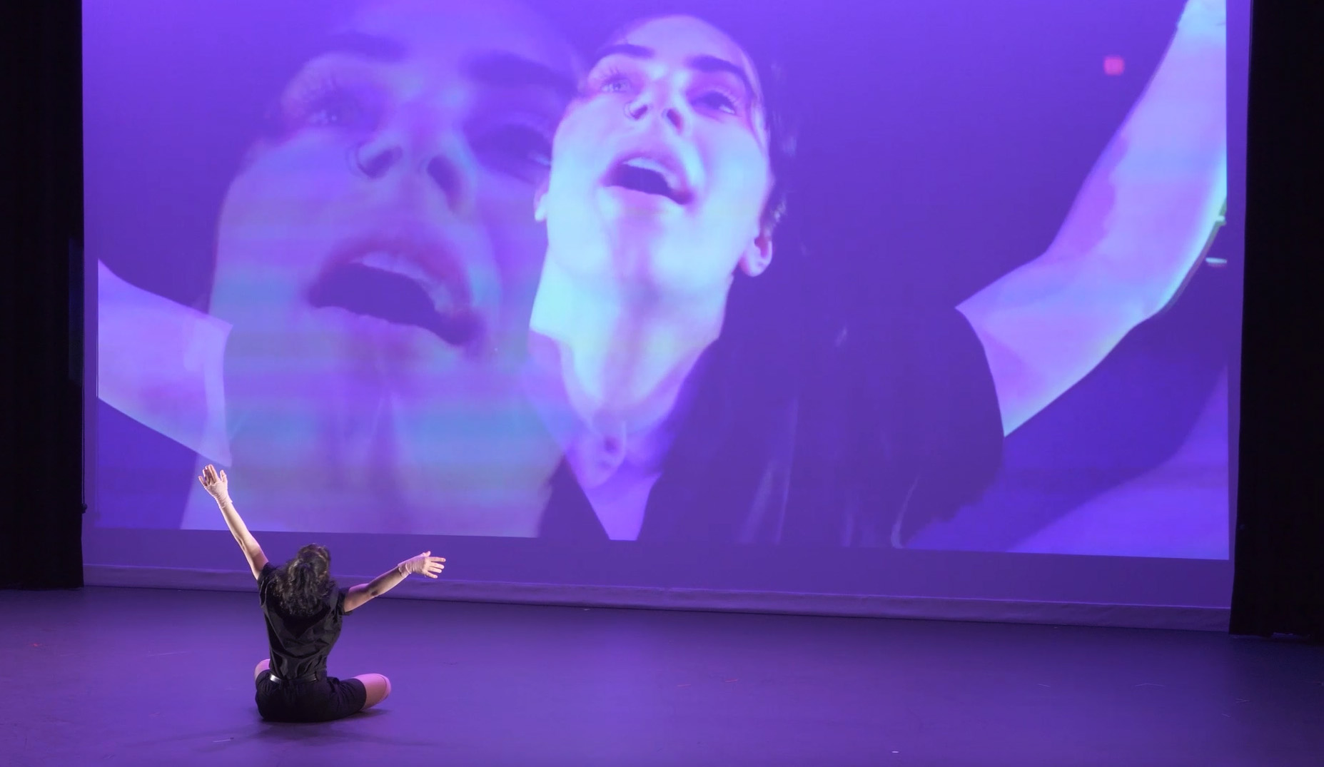 An individual sits on a stage wearing black with raised arms, facing a large screen image of a magnified reflection of themself.