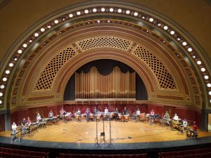 A semi-circle of 18 masked performers pose in front of assorted percussion instruments on stage at Hill Auditorium.