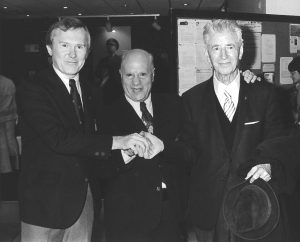 Three men in suits pose standing, linked in a three-way handshake. Black and white photo.