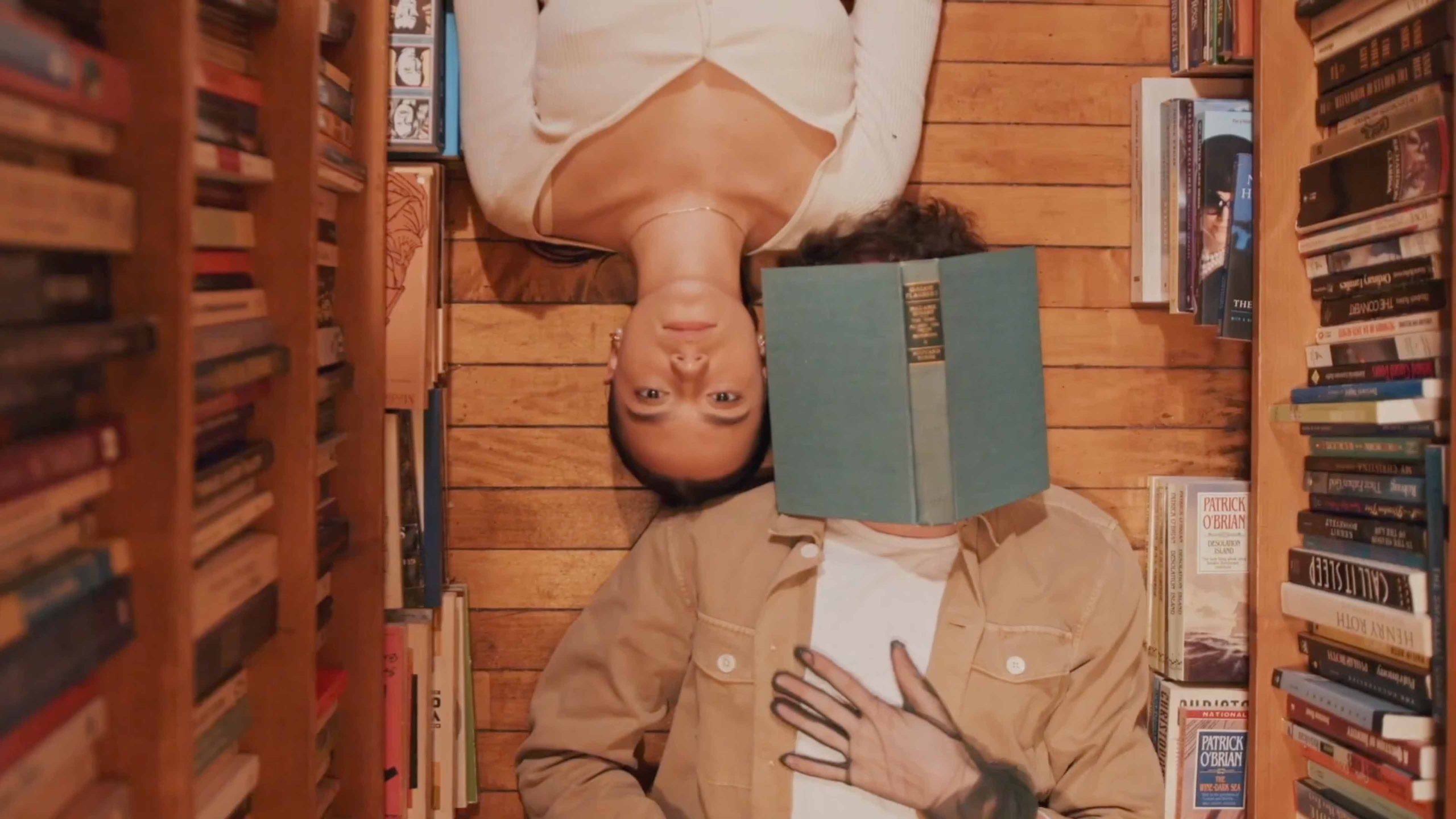 Two people laying down in the aisle of a book store; a book covers one of their faces.