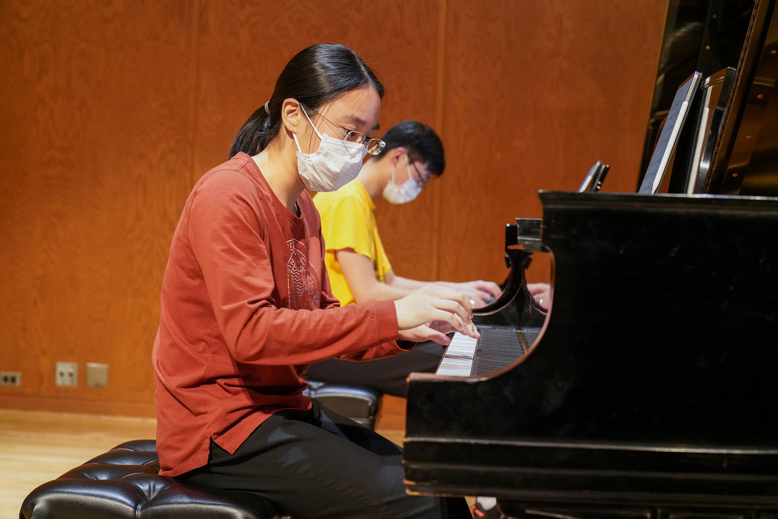 Two masked players sit together playing the same piano.