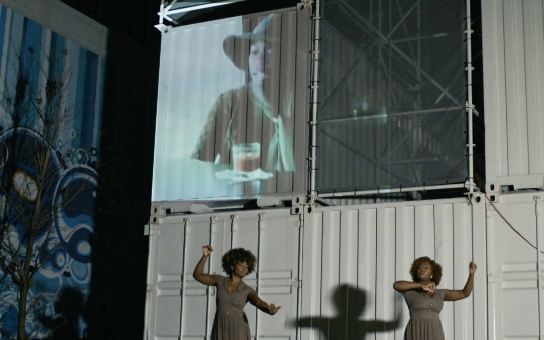 Two dancers dressed alike cast shadows in front of a mural and screen projection.