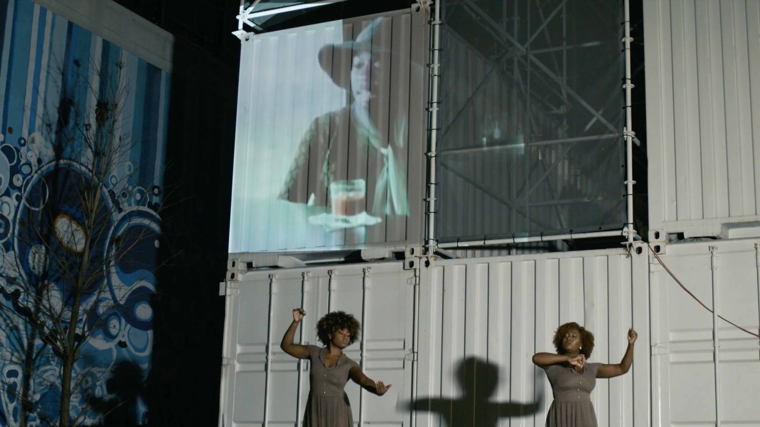 Two dancers dressed alike cast shadows in front of a mural and screen projection.
