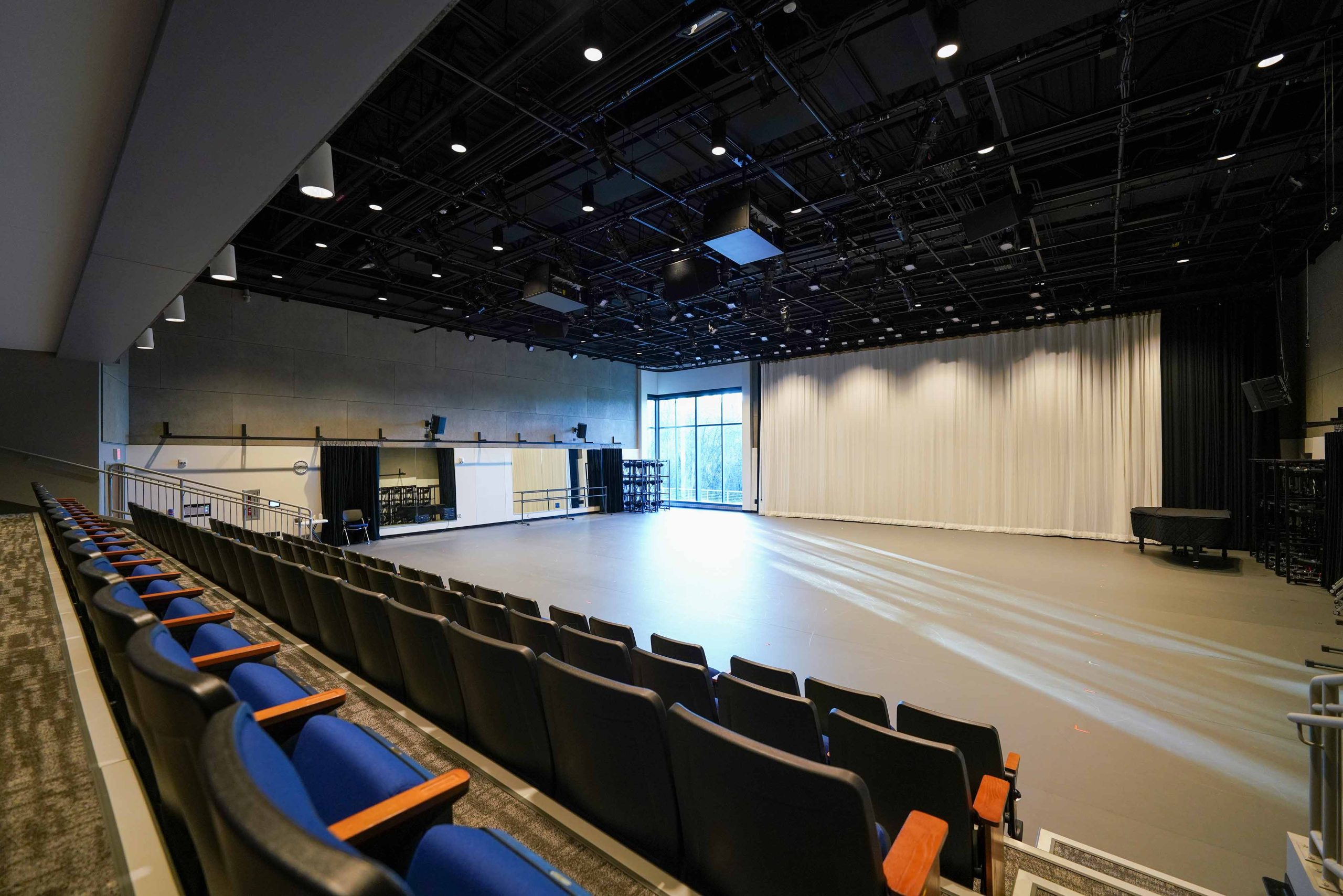 Two rows of seating overlook a studio with tall windows partially covered by white curtains.
