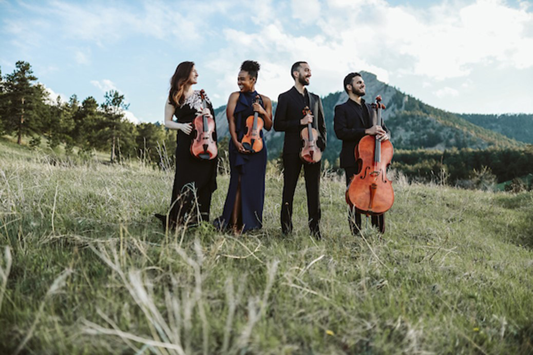 Four string players hold their instruments standing in a scenic mountain valley.