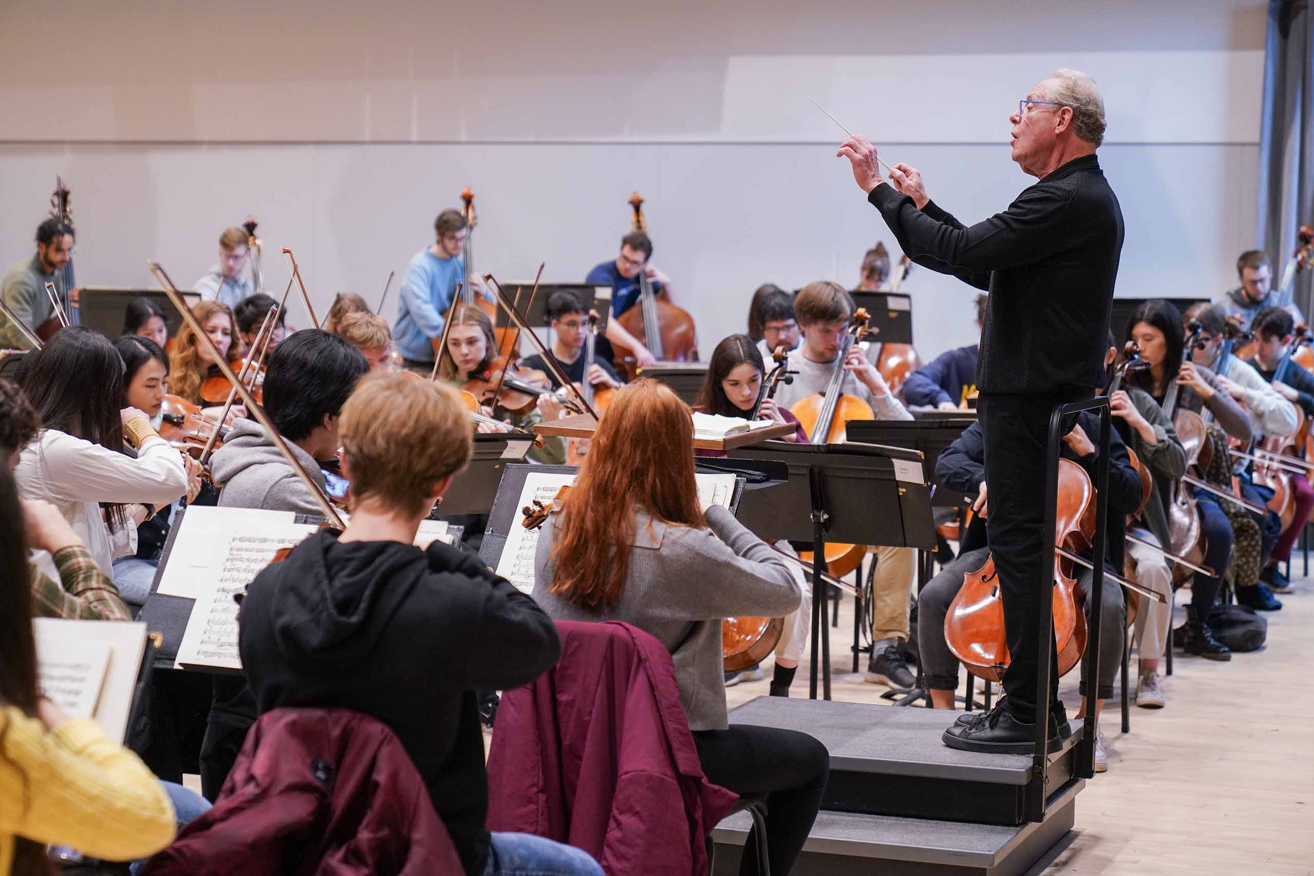 A conductor wearing all black leads an orchestra playing in a classroom