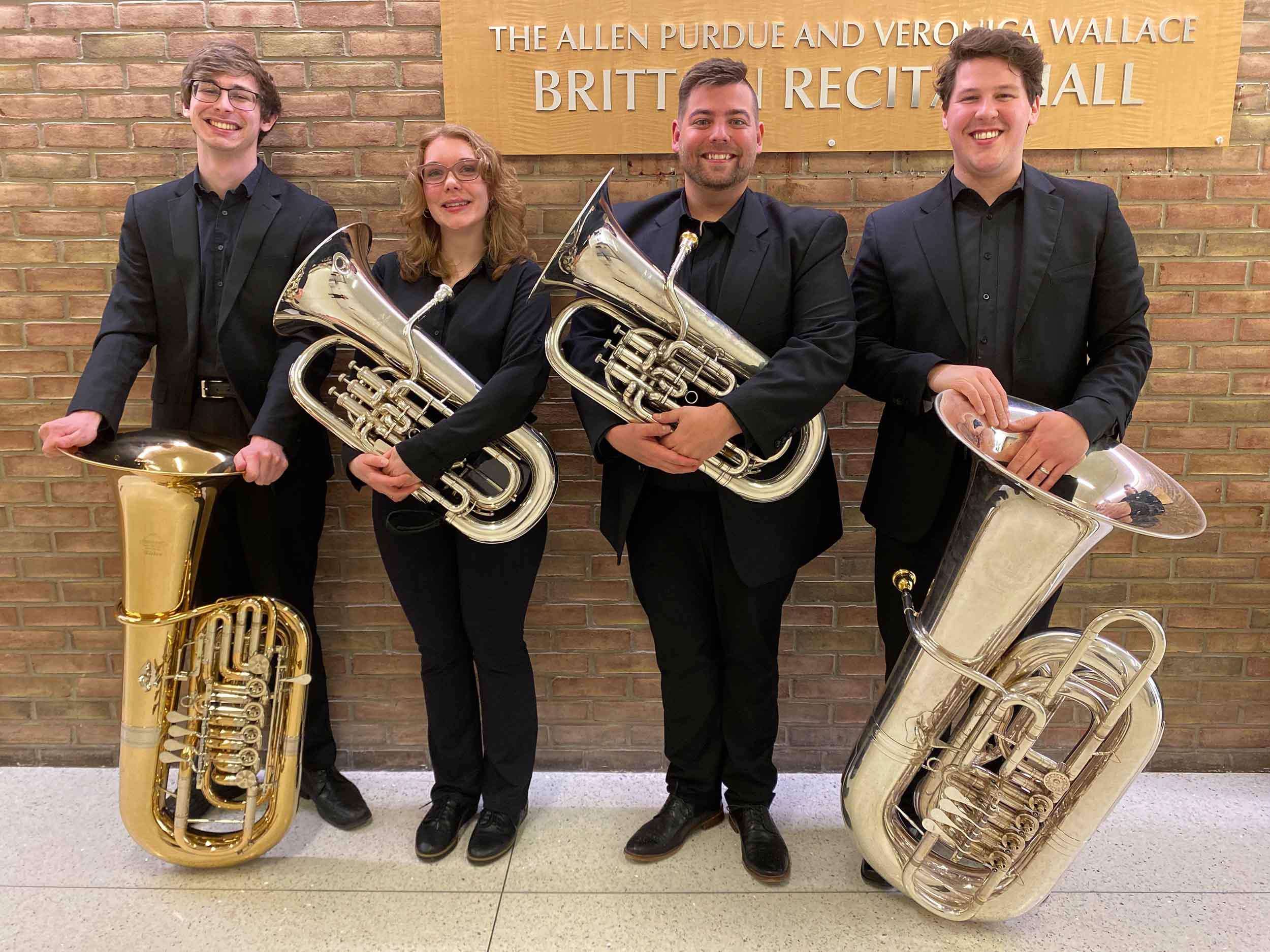 Four musicians pose standing wearing black; each holding a tuba