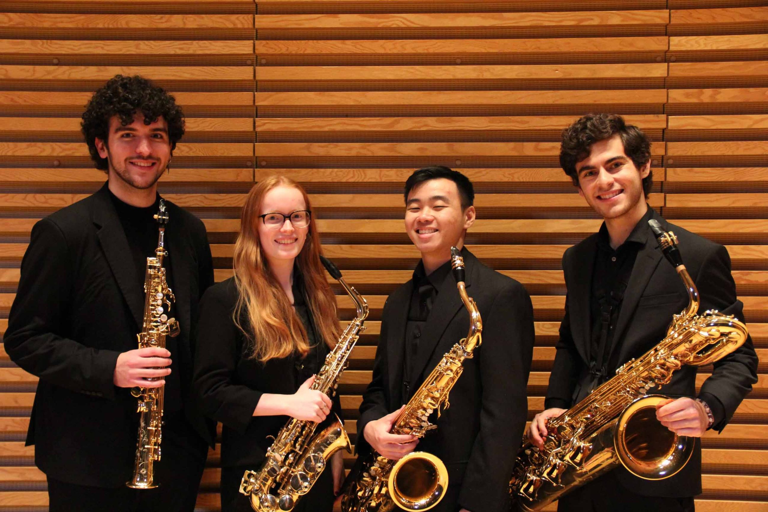 Four saxophonists wearing black stand posing, holding their instruments