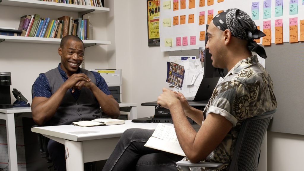 Michael McElroy and Sevon Askew sit acros from one another over a desk engaged in conversation