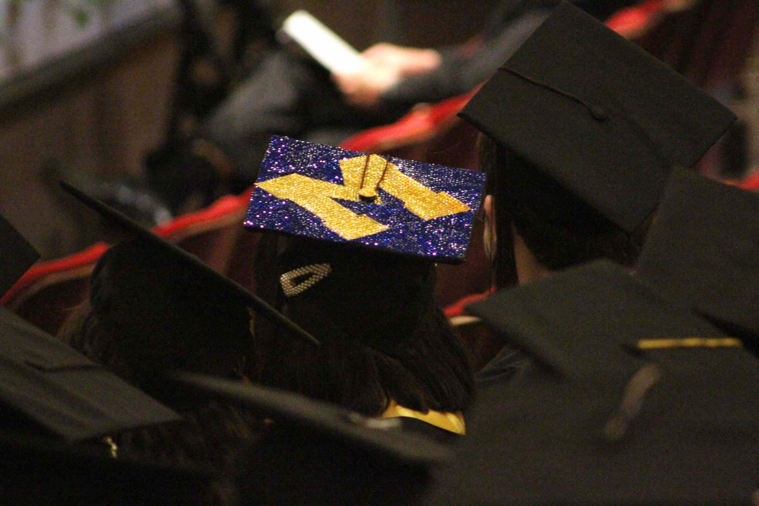 Amid rows of black mortar board graduation caps, one is covered in a glittering block M.