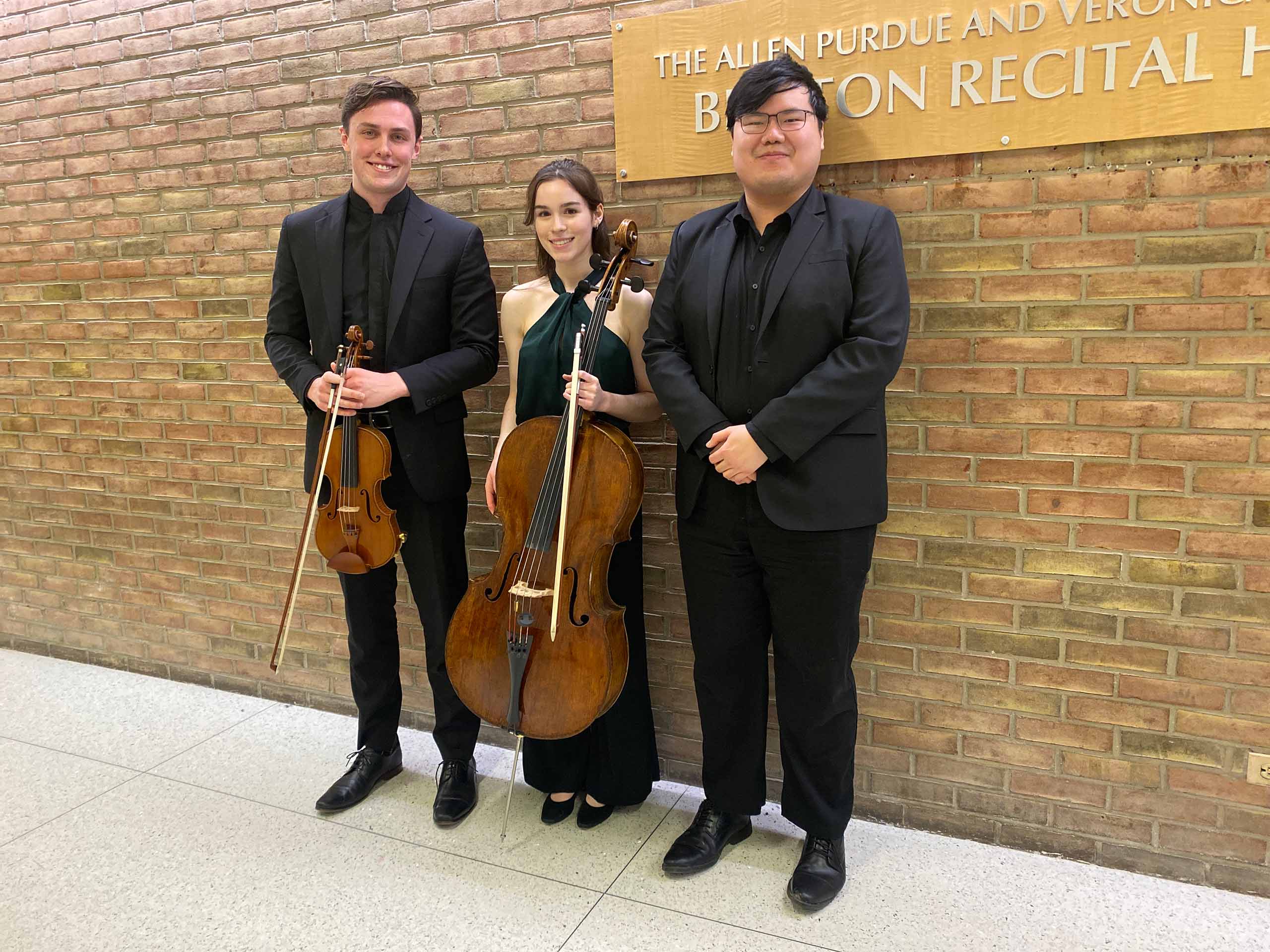 Three musicians pose standing wearing black, the first two hold a violin and a cello
