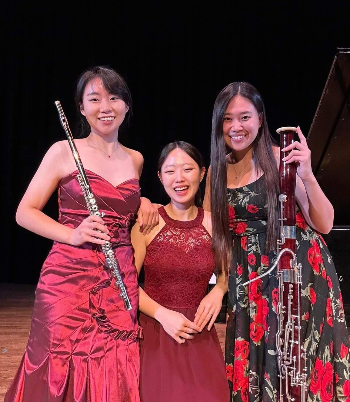 Three musicians pose on stage wearing fancy dresses; the first holds a flute and the third holds a bassoon