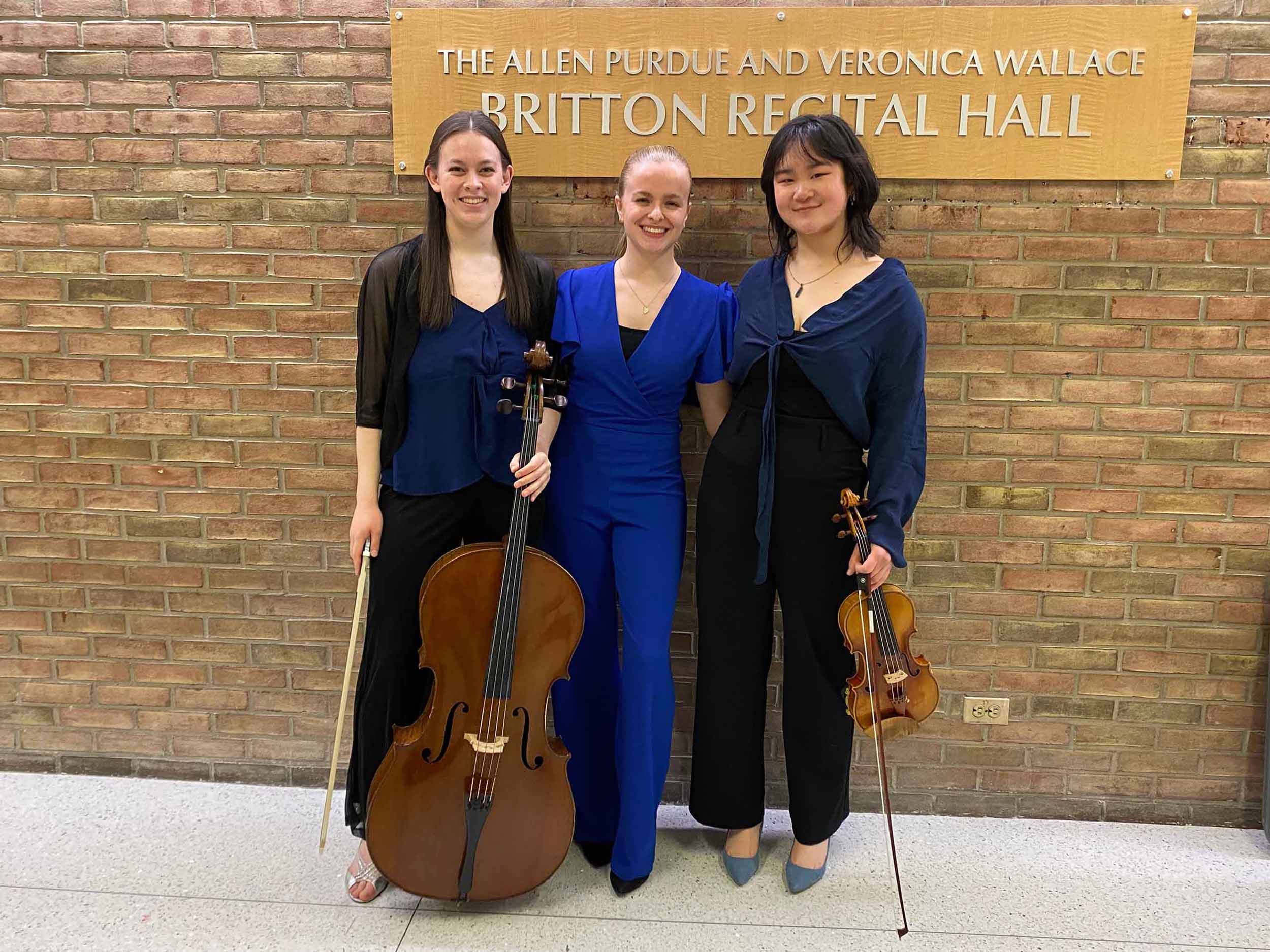 Three musicians pose standing wearing blue; the first holds a cello and the third holds a violin