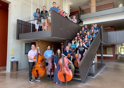 25 individuals holding string instruments pose on the lobby staircase in Walgreen Drama Center.