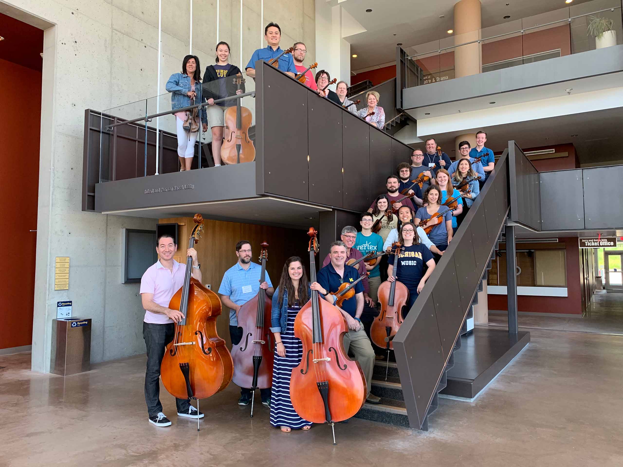 25 individuals holding string instruments pose on the lobby staircase in Walgreen Drama Center.