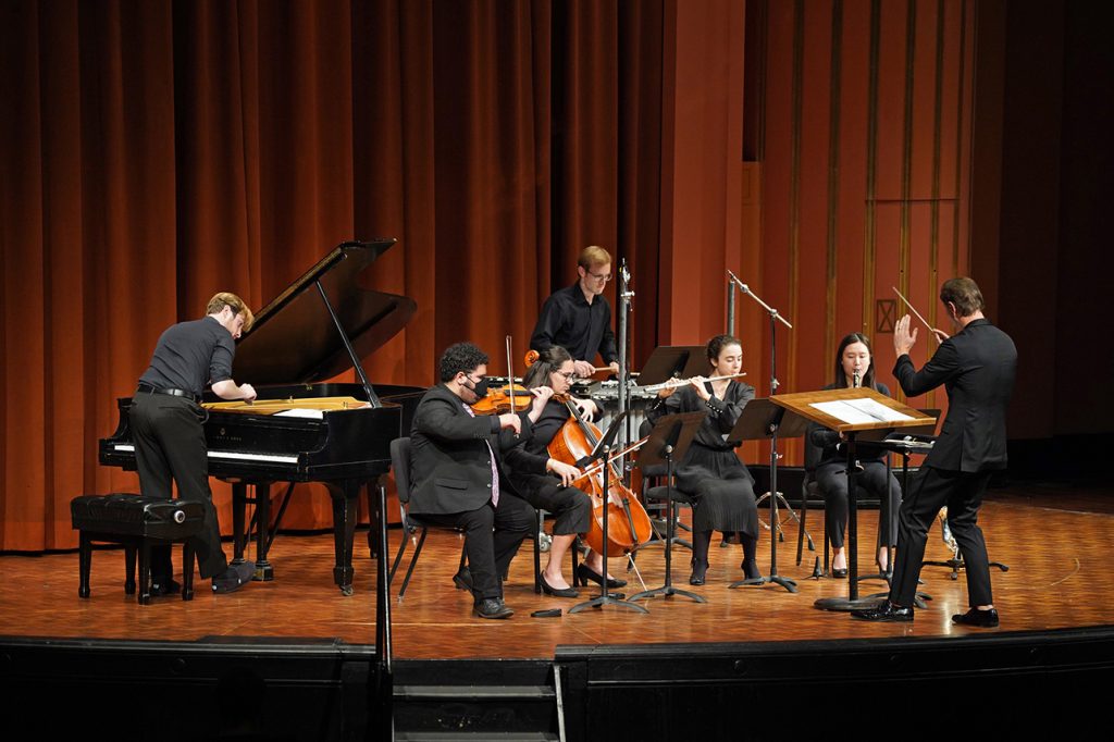 An ensemble of six players on strings, winds, percussion, and piano perform on stage, led by a conductor