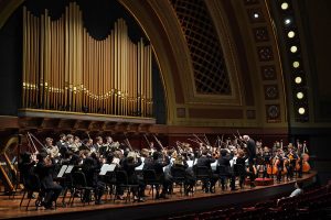 The conductor and string players hold their arms raised as the University Symphony Orchestra performs on stage at Hill Auditorium