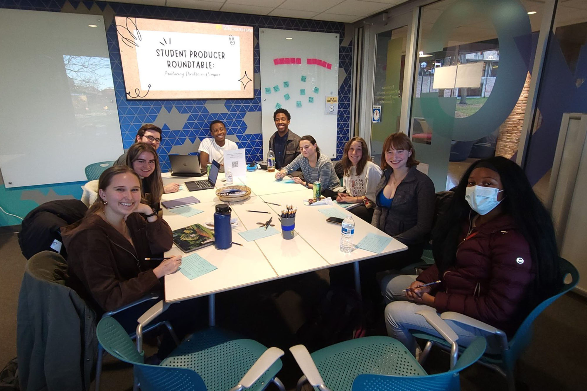 Students gathered in the EXCEL Lab around a conference table. In the background is a screen with the title "Student Producer Roundtable"