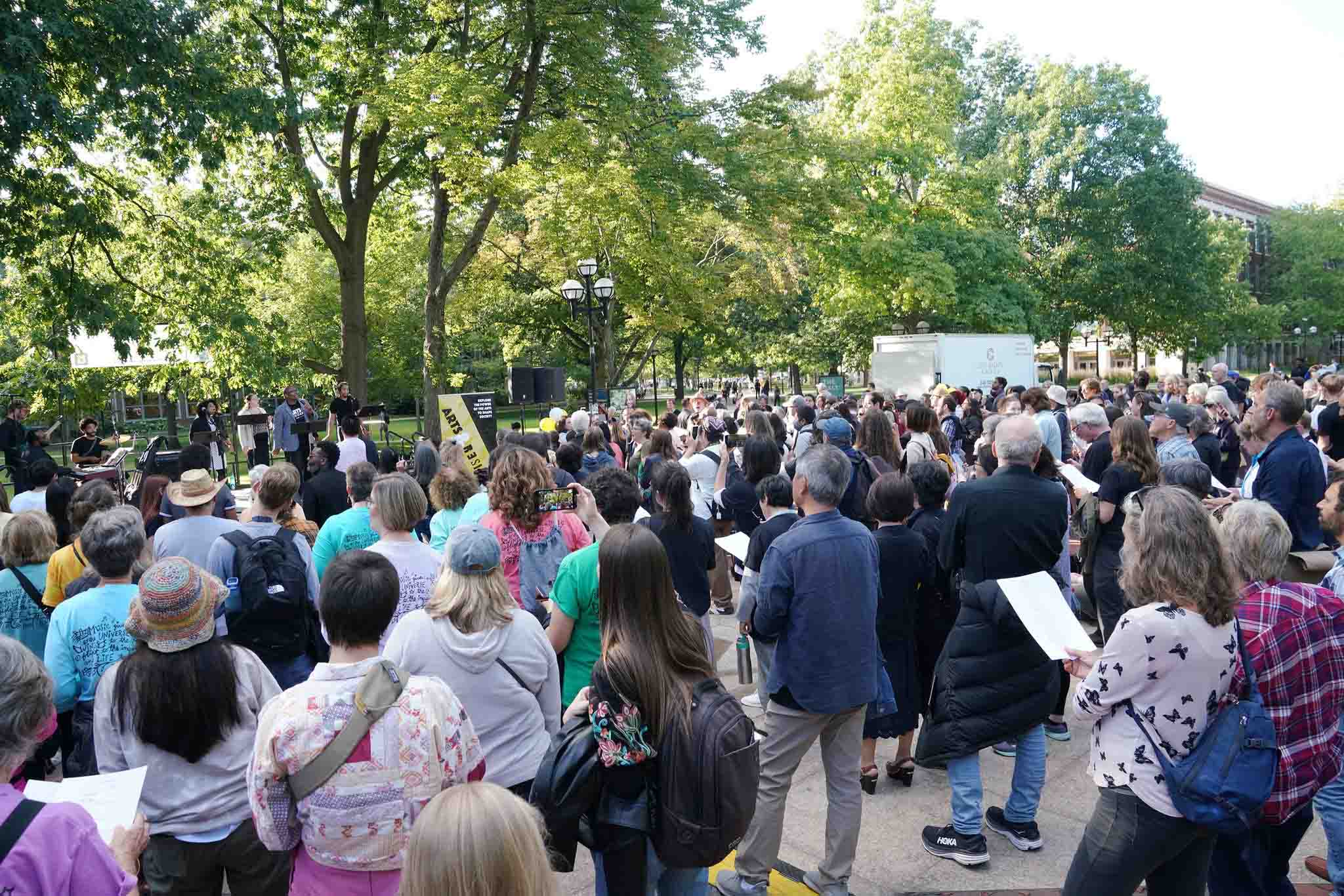 Crowd of people faces a stage on the Diag, some holding sheet music.