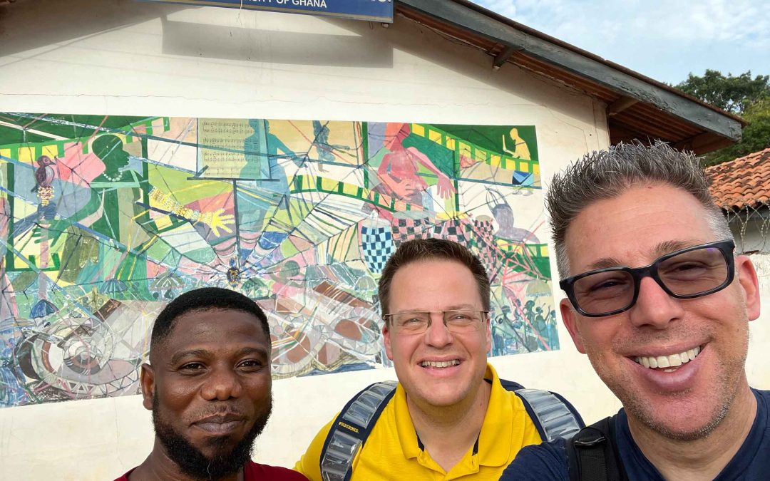 Three men smile in front of a colorful mural on a building marked "School of Performing Arts - University of Ghana"