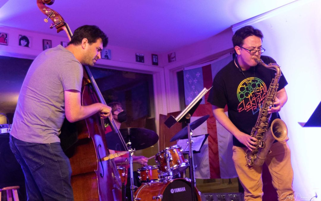 Trio of bass, drums, and saxophone perform with purple backlighting