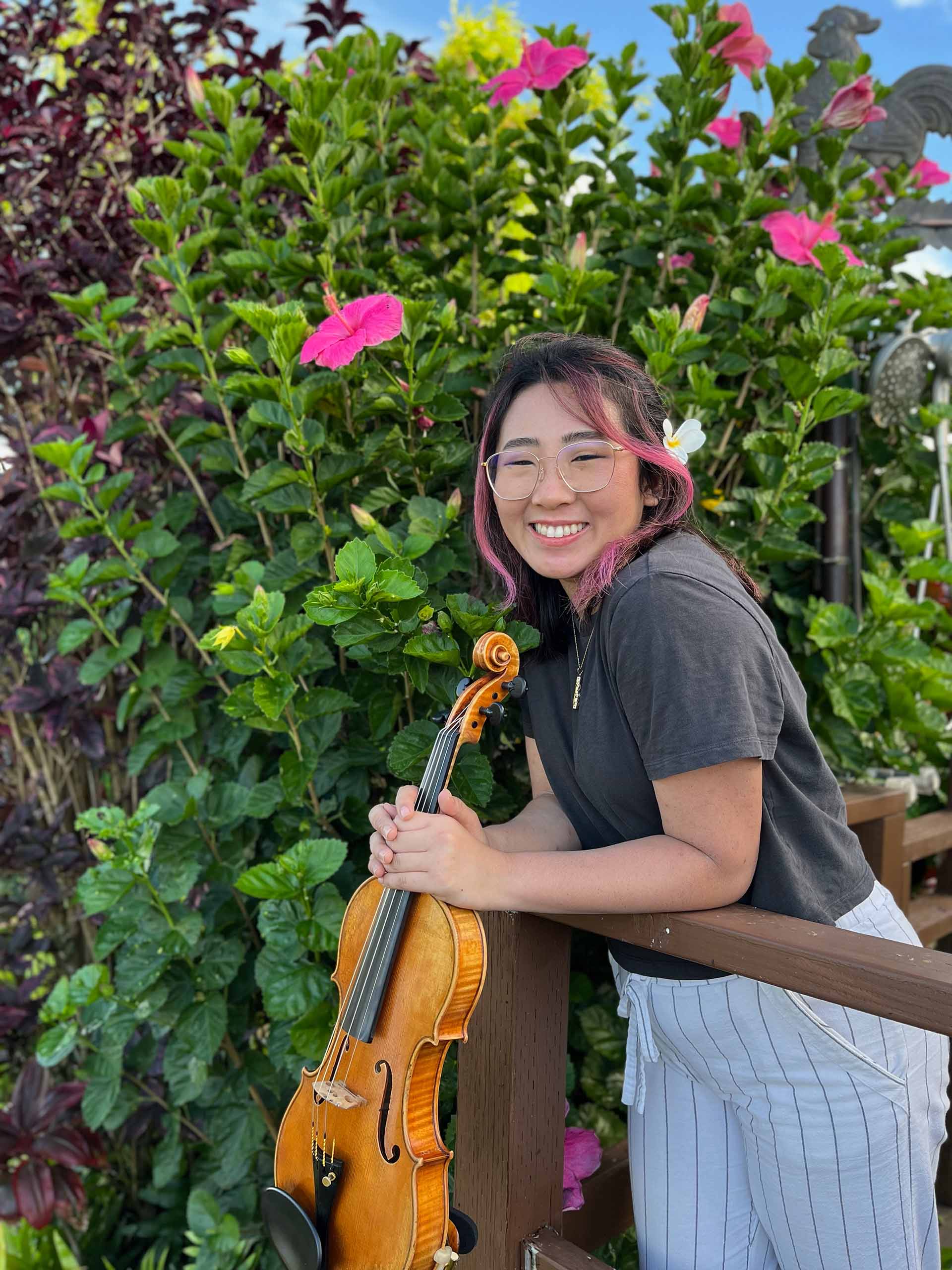 Kisa Uradomo holds her violin in front of bright pink hibiscus flowers