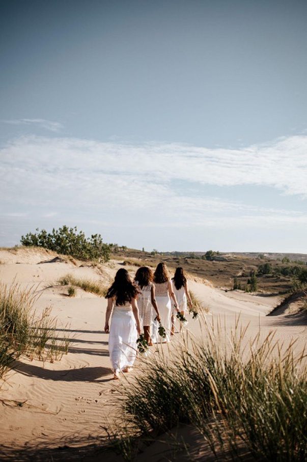 Four figures wearing white dresses walk away in a line, holding flower bouquets, in a vast dunescape
