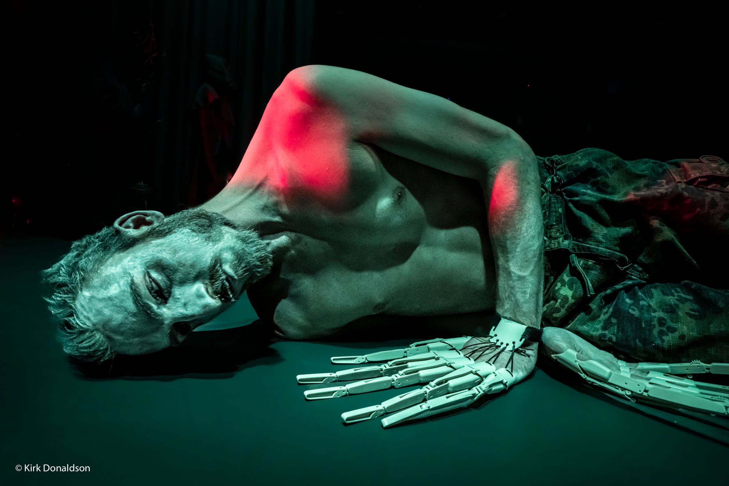 A man lays wearing two robotic hands, green and red lighting cast on him